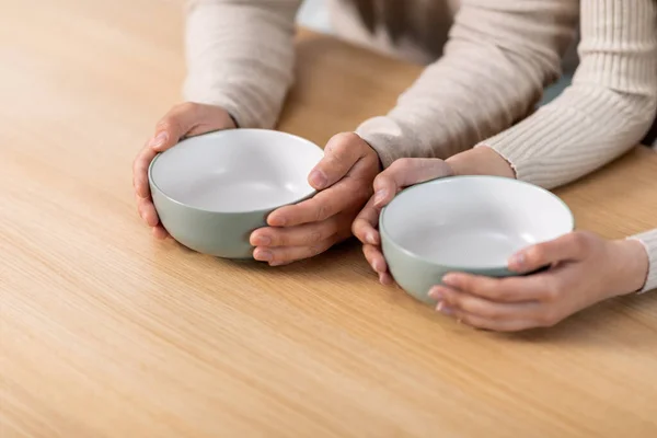 Unrecognizable people husband and wife sitting at kitchen table with spoons in empty bowls, poor hungry family has nothning to eat, suffering from hunger while crisis during pandemic, cropped