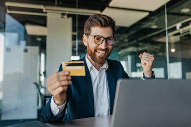 Happy businessman holding credit card in front of laptop and making YES gesture, shopping while sitting in office interior. Cool online promo concept