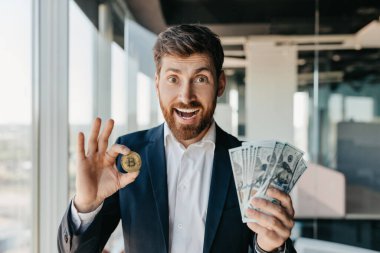 Crypto trading. Excited businessman holding golden bitcoin coin and dollar cash banknotes, happy male trader in suit standing in modern office interior