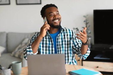 Cep telefonuyla konuşan mutlu siyahi adam dizüstü bilgisayarını kullanarak çalışıyor ve evdeki masada otururken interneti inceliyor. Çevrimiçi İş ve Mobil İletişim Konsepti. Seçici Odaklanma