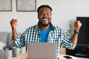 Dizüstü bilgisayar kullanan neşeli siyah erkek ve evde oturan kameraya gülümseyen yumruklar sallıyor. Serbest Çalışan Adam İş Başarısını Kutluyor. Başarılı Serbest Kariyer