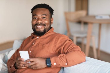 Neşeli Siyah Erkek, yeni uygulamasını cep telefonunda kullanıyor. Evdeki koltukta oturan kameraya gülümsüyor. Oturma odasında akıllı telefonlu sakallı adam pozu. Mobil Teknoloji Konsepti