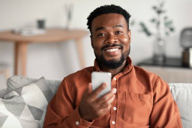 Cheerful African American Guy Using Mobile Phone Advertising New Application Sitting On Couch Smiling To Camera At Home. Technology, Great App Concept. Selective Focus