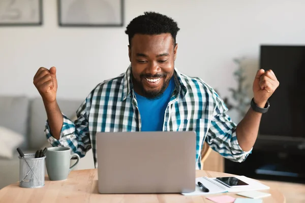 Joyful African American Guy Using Laptop Shaking Fists Celebrating Success And Great Online News Sitting At Home. Freelancer Man Gesturing Yes Working Remotely Indoor