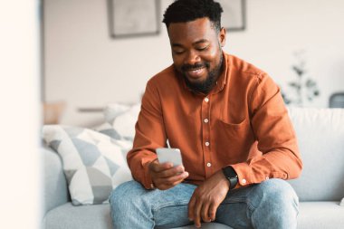 Mutlu Siyahi Adam Cep Telefonu ile İnternet 'i tarıyor ve evdeki koltukta oturup mesajlaşıyor. Guy Mobil Uygulama Web Sörfü ve Ağ Kullanıyor. Özellikler Yaşam Biçimi Konsepti