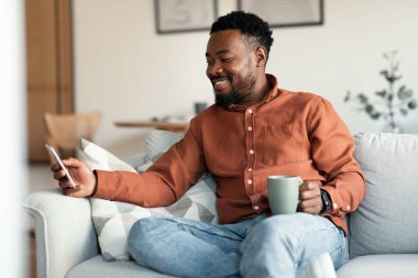 Smartphone 'da uygulamayı kullanıp evdeki koltukta oturup kahve içen mutlu siyah adam. Hafta sonu cep telefonuyla erkek iletişimi mesajlaşması. Mobil İletişim ve Teknoloji