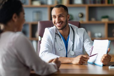 Cheerful young arabic general practitioner showing black lady patient treatment plan, african american woman attending therapist at newest private clinic, modern healthcare concept