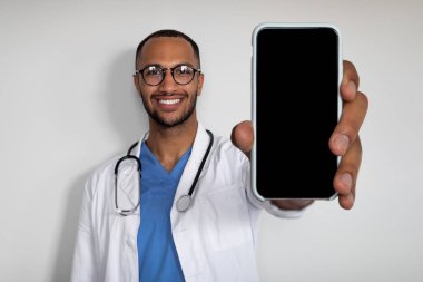 Handsome young arabic man practitioner showing newest smartphone with black empty screen and smiling, recommending brand new application for healthcare, white background, mockup