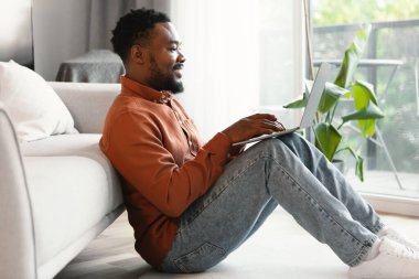 Siyah erkek serbest bilgisayar kullanıyor, bilgisayarda çalışıyor ve internette geziniyor, evde oturuyor, kenar manzaralı. İnternet sörfü yapan adam uzaktan iş yapıyor. Serbest ve Teknoloji Konsepti