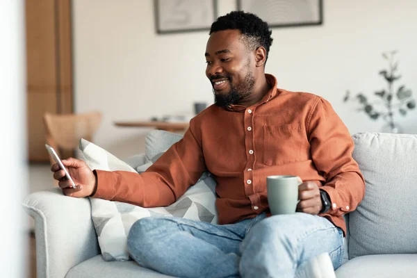 Hombre Negro Feliz Usando Aplicación Smartphone Beber Café Sentado Sofá — Foto de Stock