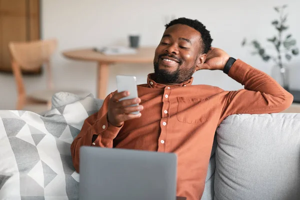 Cheerful African American Guy Using Smartphone Laptop Browsing Internet Sitting —  Fotos de Stock