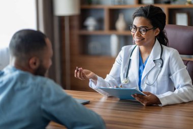 Cheerful pretty african american young woman doctor taking anamnesis of millennial middle eastern guy patient, sitting together at workdesk, therapist holding medical chart. modern clinic interior