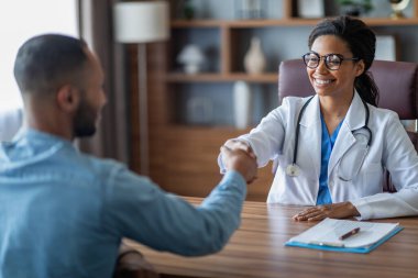 Friendly young african american lady doctor smiling and shaking male patient hand, cheerful black woman therapist greeting middle eastern guy at her clinic, outstretching hand, copy space
