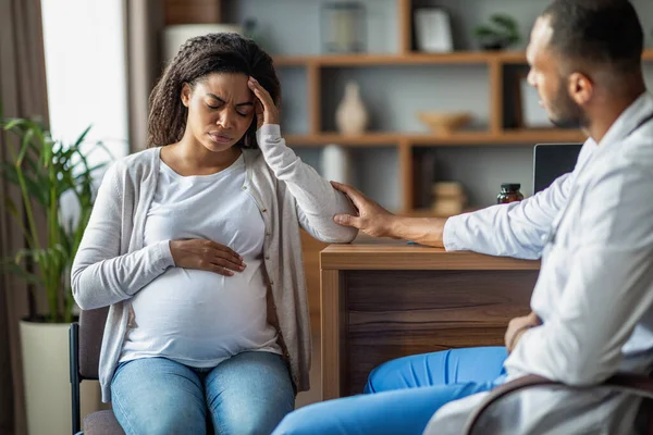 Pregnant millennial black woman suffering from migraine, sitting on chair at clinic, touching her big tummy and head, visiting doctor, arab man gynecologist comforting his expecting patient