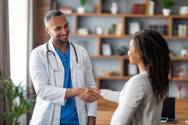 Handsome young arabic doctor gynecologist smiling and shaking female patient hand, cheerful middle eastern man therapist greeting pregnant african american lady at his clinic, outstretching hand