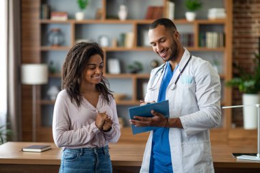 Friendly handsome arab man doctor consulting young african american lady patient, doc giving recommendations, looking at medical chart, standing next to workdesk at modern clinic