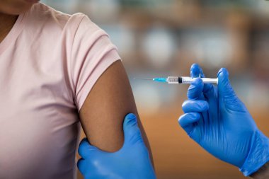 Doktor ya da hemşire ellerinde mavi eldivenlerle siyah kadın hastaya iğne yapıyor. Afro-amerikan bayanın COVID ya da grip aşısı oluyor. Geçmişi bulanık.