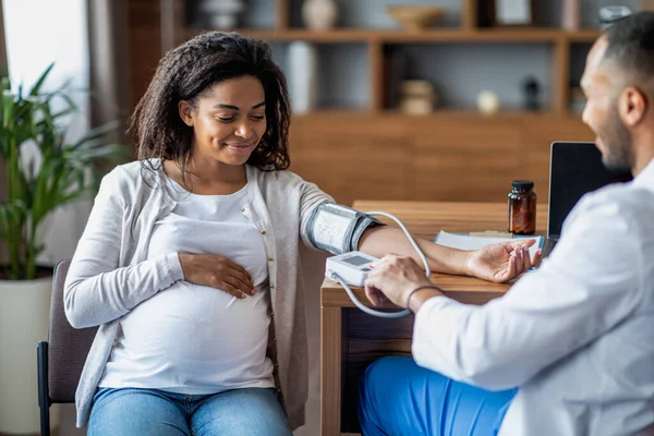 Middle eastern man doctor gynecologist checking happy pregnant african american woman heart rate and blood pressure, using tonometer, clinic interior, copy space. Medical care during pregnancy