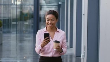 Young african american business lady walking with takeout coffee and smartphone near office building, spending her lunch break outdoors, follow shot, slow motion