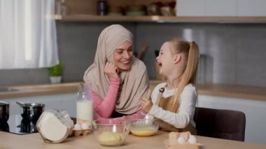 Cooking is fun. Adorable little girl whisking eggs in bowl for breakfast and laughing, loving muslim mother wearing hijab watching her happy child at kitchen