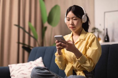 Cep telefonu kullanan Asyalı bir kadın kablosuz kulaklık takıyor Audiobook 'un Evdeki Koltukta Oturarak Öğrenmesini dinliyor. Milenyum Kadını Akıllı Telefon 'da Müzikal Uygulama Kullanıyor