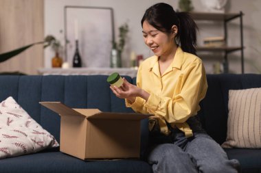 Mutlu Koreli Bayan Kozmetik Ürünleri Teslimatı Yapıyor Nemlendirici Kavanozu Evdeki koltukta karton kutunun yanında oturuyor. Kozmetoloji, Başarılı Makyaj Alışveriş ve Teslimat Konsepti