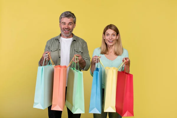 Big sale and shopping with pleasure. Excited middle aged spouses holding many colored packages in hands, posing on yellow background, studio shot, copy space