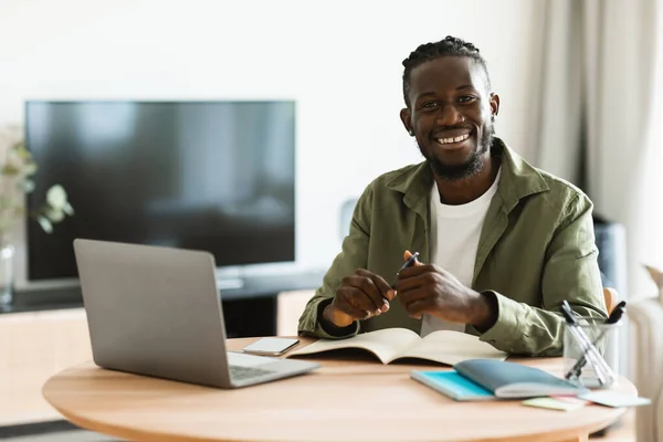 Portrait of african american freelancer guy sitting at desk at home office, looking at camera and smiling, free space. Black man enjoying remote work and self-employment