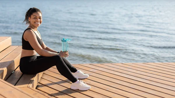 Sport and hydration. Happy millennial black woman jogging by seaside alone, drinking water while having break, looking at camera and smiling, panorama, with copy space, full length shot