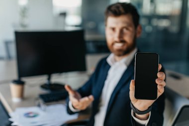 Mobile offer. Businessman showing blank smartphone while sitting at desk in office interior, selective focus. Entrepreneur demonstrating space for advertisement ar app design, mockup
