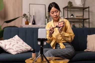 Japon blogcu kadın cep telefonuyla video çekiyor kozmetik malzemeler kullanarak evdeki koltukta oturuyor. Kadın Reklamcılık Kozmetik. Profesyonel Güzellik Blog 'u