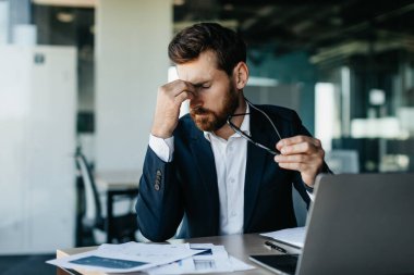 Exhausted male manager or businessman sitting in front of laptop at modern office interior, holding glasses and rubbing nose bridge, having headache