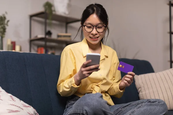 Mobile Shopping. Japanese Lady Using Cellphone And Credit Card Making Payment Sitting On Couch At Home. Ecommerce, Mobile Banking Application Concept. Selective Focus