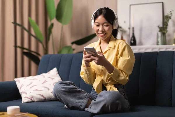 Cheerful Japanese Millennial Woman Using Cellphone Wearing Wireless Earphones Sitting On Couch At Home. Lady Using Musical Application And Browsing Internet. Gadget Lifestyle