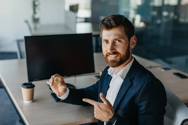 Happy businessman in suit holding golden bitcoin and pointing finger on it, sitting at workplace with computer, trading on stocks and markets, working at office. Cryptocurrency concept