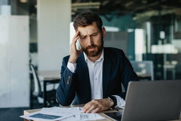 Stressed entrepreneur suffering headache, sitting at desk with lot of papers, tired businessman having acute migraine, rubbing temples and frowning, office interior