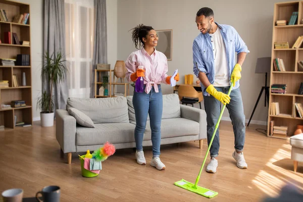 House Chores. Cheerful Black Spouses Cleaning Living Room Doing Housework Together Standing At Home. Husband And Wife Holding Mop And Detergent Bottle. Full Length Shot