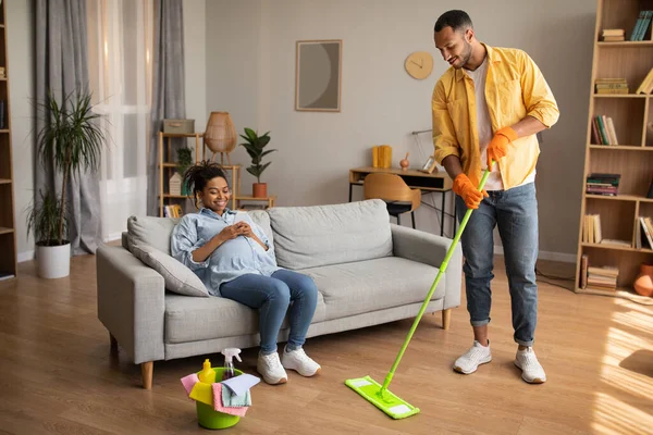 Black Husband Doing House Chores Cleaning Living Room While Pregnant Wife Resting Sitting On Couch At Home. Pregnancy And Family Lifestyle, Leisure Concept. Full Length Shot