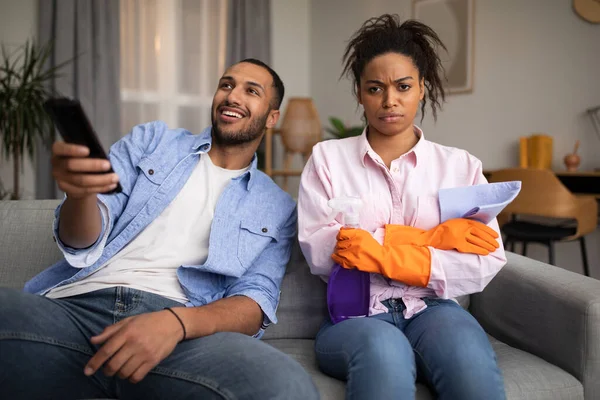 Nespokojeni Black Millennial Manželka Úklid Dům Zatímco Lenivý Manžel Sledování — Stock fotografie