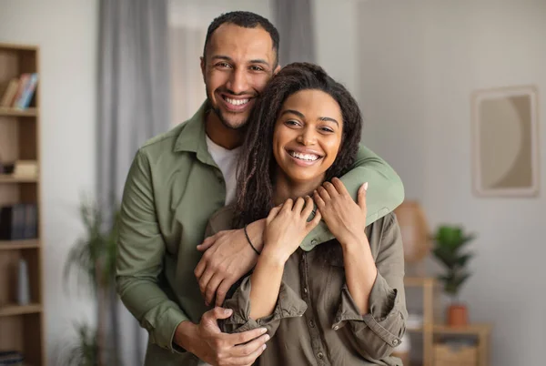 Happy Marriage. Joyful African American Husband Hugging Wife Smiling Looking At Camera Standing At Home. Happy Couple Embracing Posing Together. Love And Romantic Relationship Concept