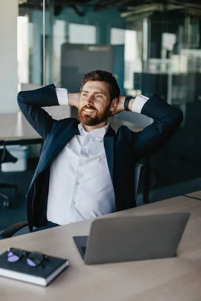 Taking break from work. Excited businessman relaxing on chair in his office, leaning back at workplace and holding hands behind head, looking away and smiling