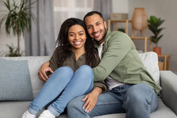 Loving African American Couple Embracing Posing Smiling To Camera Sitting On Sofa At Home. Cheerful Boyfriend Hugging Girlfriend Spending Time Together. Happy Romantic Relationship