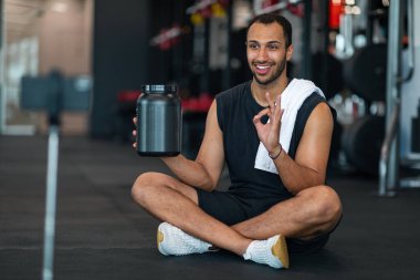 Spor salonunda Whey Powder için video incelemesi yapan yakışıklı siyah erkek atlet, Afrikalı Amerikalı spor blogcusu sosyal medyada yeni spor ürünü sunuyor, üç ayaklı araçta akıllı telefon kullanıyor.
