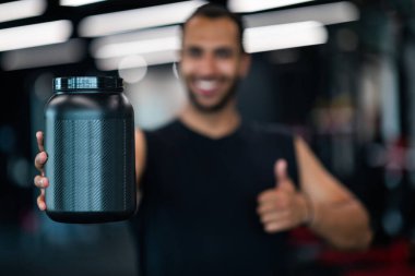 Fitness Nutrition. Beyaz Protein tozu ve kameraya el hareketi yapan, gülümseyen Afrikalı Amerikalı erkek sporcu Yeni Spor Beslenme Ürünleri Öneren Mutlu Siyah Sporcu