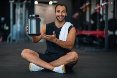 Gülümseyen Sporcu Siyah Adam Kamerada Beyaz Protein tozuyla konteynır gösteriyor, Afrikalı Amerikalı erkek sporcu spor malzemeleri Modern Spor Salonu 'nda poz verirken, Uzay Kopyalama