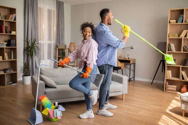 Happy African American Couple Doing Housework Cleaning House Together And Singing Having Fun Posing With Broom And Mop Standing In Modern Living Room At Home. Full Length