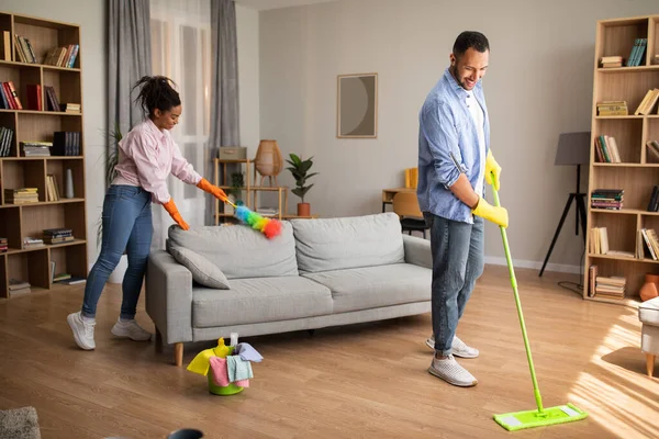 House Chores. African American Husband And Wife Cleaning Modern Living Room With Mop And Feather Duster Standing At Home On Weekend. Spouses Doing Housework Together
