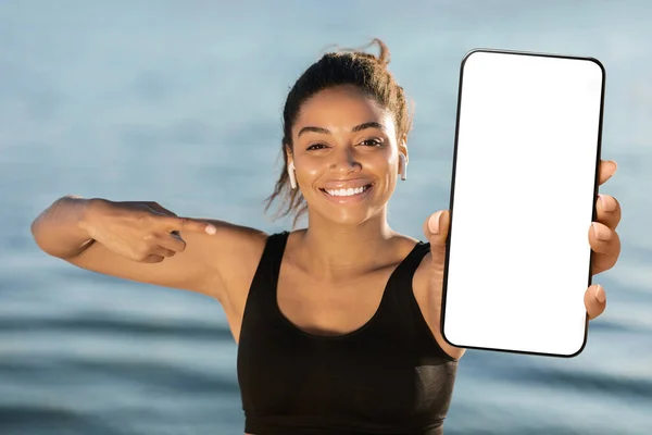Happy attractive athletic millennial black woman pointing at brand new smartphone with white empty screen and smiling, recommending newest mobile fitness app, mockup, seaside background