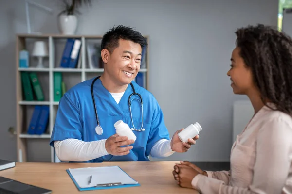 Happy young asian man doctor in uniform consults to black lady patient, offers jars with pills in office clinic interior. Health care, meeting with therapist and treat disease and medicines, pharmacy