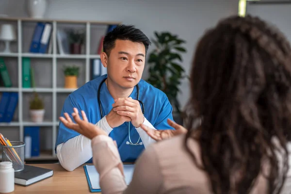 Serious Millennial Chinese Guy Doctor Uniform Listens Symptoms African American — ストック写真